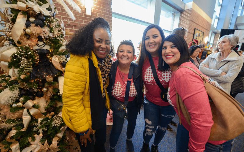 Group of four 40-something smiling women at  KC Holiday Boutique in line beside Christmas Tree