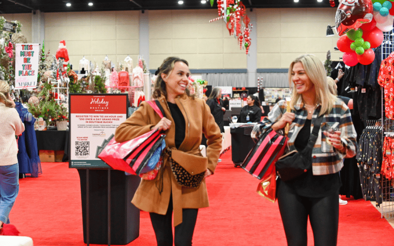 Two young women walking down red carpet on show floor at KC Holiday Boutique smiling at each other and carrying shopping bags 
