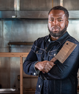 Ryan Mitchell with Meat Cleaver wearing navy blue denim jacket posing in industrial kitchen