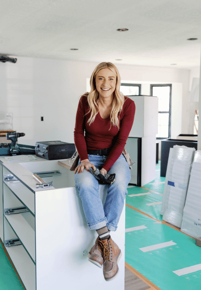 Jasmine Roth wearing long sleeved red top and jeans sitting on counter in room under renovations